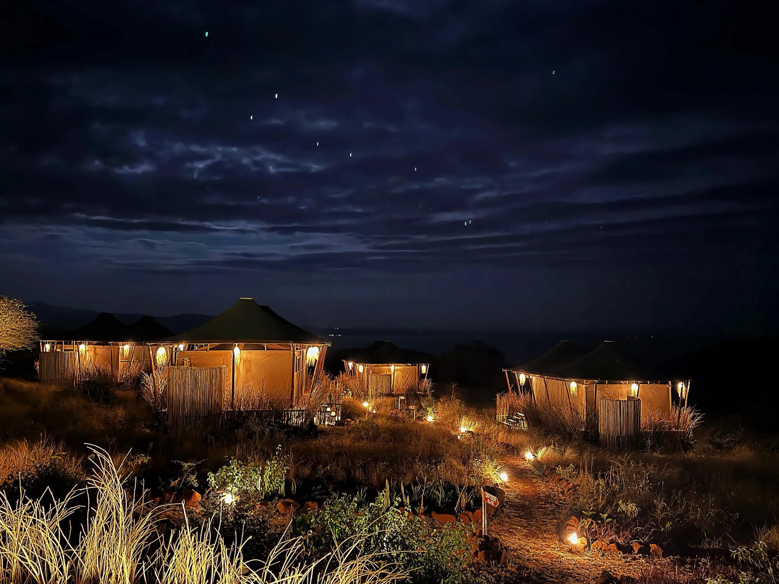 The camp after dark — lanterns lit along the path between the tents, Lake Manyara beyond